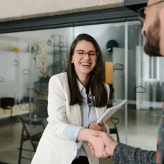 Uśmiechnięta kobieta w okularach i marynarce ściska dłoń mężczyźnie. Może to być początek nowej ścieżki kariery, choć częsta zmiana pracy nie zawsze jest łatwa.