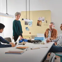 Zespół dyskutuje przy stole, inspirując się nawzajem. Kobieta z siwymi włosami, uśmiechnięta, uosabia przywództwo transformacyjne, motywując do kreatywności.