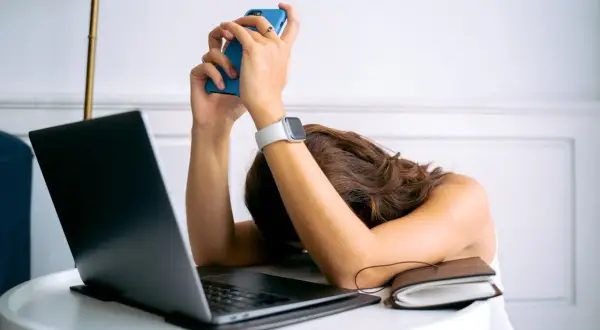 Kobieta z opuszczoną głową na biurku, trzymająca telefon. Wokół laptop i notatnik. Czuje, że ma dość wszystkiego, cytaty z życia.