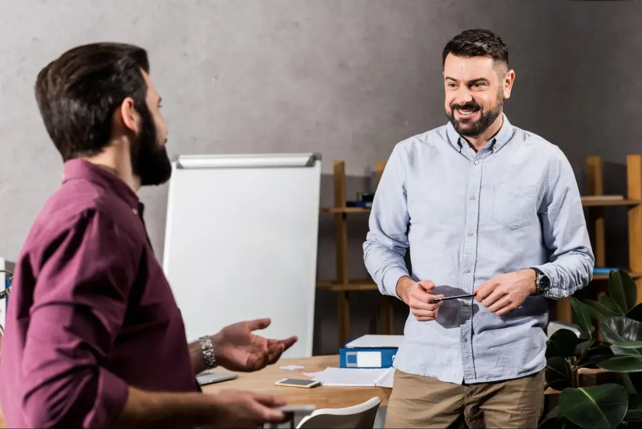 Dwóch mężczyzn rozmawia przy biurku. Jeden z nich, w niebieskiej koszuli, trzyma długopis i uśmiecha się, jakby tłumaczył, jak utemperować pracownika.